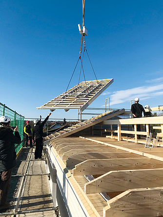 Drei Baubeschäftigte nehmen ein vorgefertigtes Dachelement in Empfang, das per Kran und Traverse von oben auf den Dachneubau herabgelassen wird.