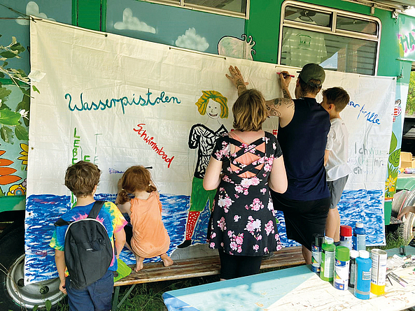 Malaktion für buntere Bauplanen am Uferstreifen Kinder und Erwachsene vor einer Malwand.