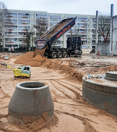 Baustelle mit Lkw beim Einbau von Sand in eine Baugrube, im Vordergrund stehen runde Schachtbauwerke aus Beton.