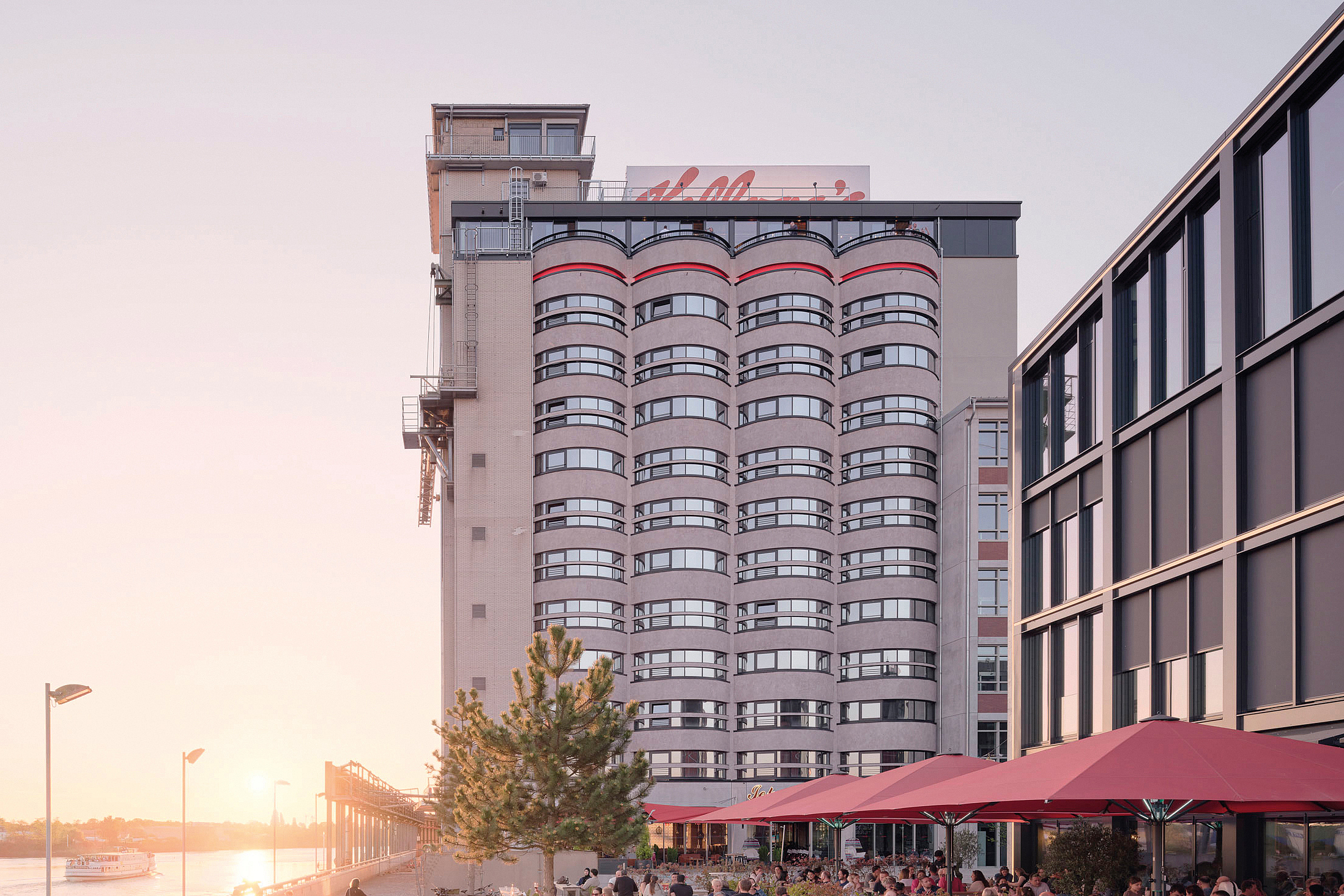 Mehrstöckiges Gebäude am Wasser mit Promenade, Außengastronomie und Menschen bei Sonnenuntergang. Auf dem Dach ist der Schriftzug "Kellogg's " angebracht.