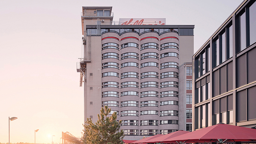 Mehrstöckiges Gebäude am Wasser mit Promenade, Außengastronomie und Menschen bei Sonnenuntergang. Auf dem Dach ist der Schriftzug "Kellogg's " angebracht.