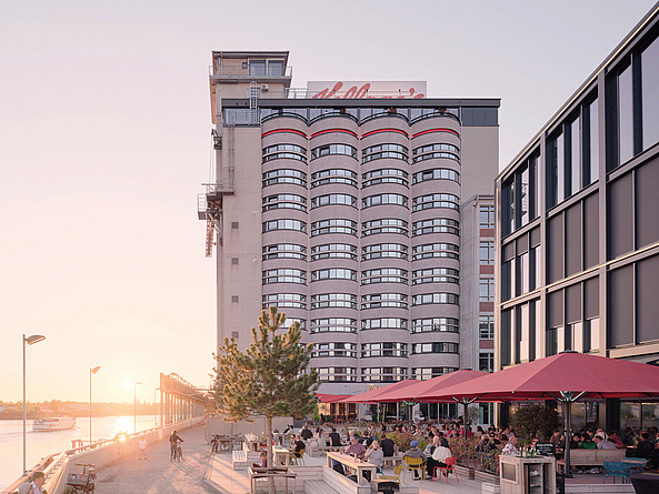 Mehrstöckiges Gebäude am Wasser mit Promenade, Außengastronomie und Menschen bei Sonnenuntergang. Auf dem Dach ist der Schriftzug "Kellogg's " angebracht.