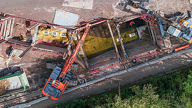 Luftaufnahme einer Baustelle mit mehreren Baugruben, einem Bagger und einem Kran. Die Arbeiten finden entlang eines länglichen Grabens mit verbauten Bauabschnitten statt.