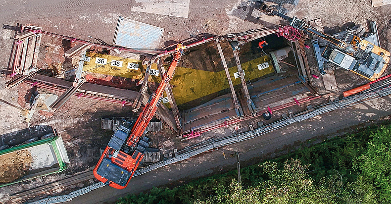 Luftaufnahme einer Baustelle mit mehreren Baugruben, einem Bagger und einem Kran. Die Arbeiten finden entlang eines länglichen Grabens mit verbauten Bauabschnitten statt.