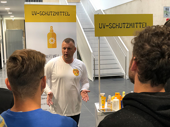 Ein Ausbilder erklärt zwei männlichen Azubis mit Plakaten und verschiedenen Sonnencremes den richtigen UV-Schutz