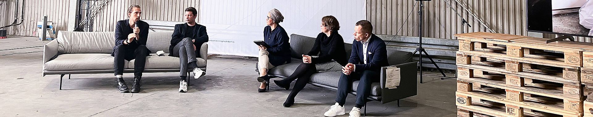 In einer Halle, die als Konferenzraum dient, sitzen fünf Personen zu einer Diskussionsrunde zusammen.