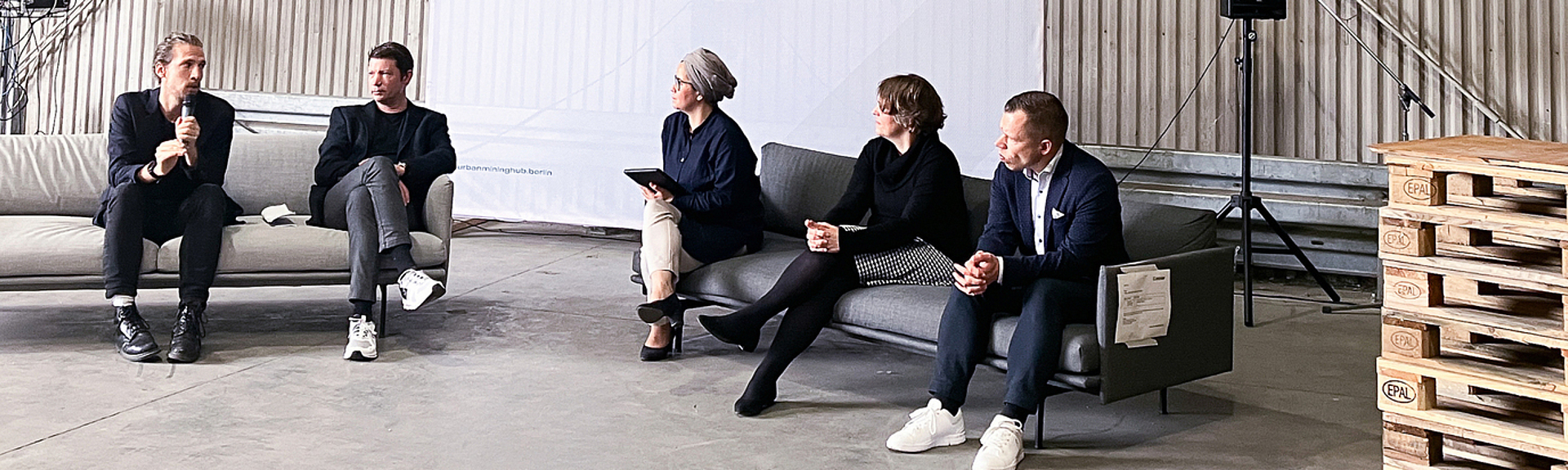 In einer Halle, die als Konferenzraum dient, sitzen fünf Personen zu einer Diskussionsrunde zusammen.