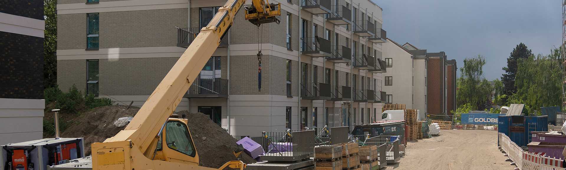 Baustelle mit mehrgeschossigem Neubau, davor befinden sich Fertigelemente und Balkone zum weiteren Anbau sowie ein Kranfahrzeug.