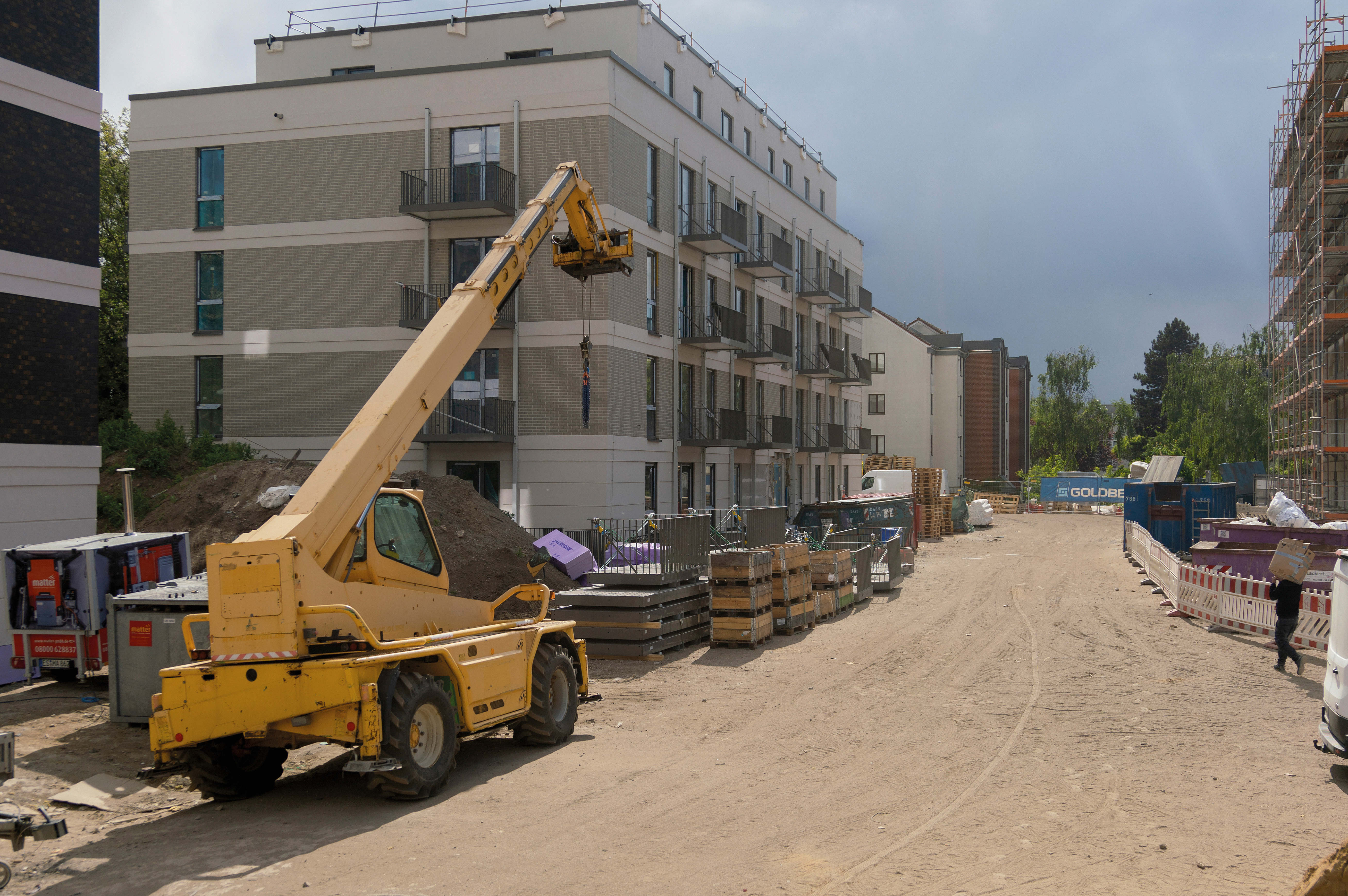 Baustelle mit mehrgeschossigem Neubau, davor befinden sich Fertigelemente und Balkone zum weiteren Anbau sowie ein Kranfahrzeug.