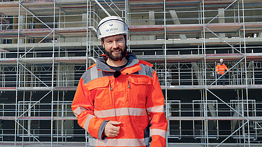 Eine Aufsichtsperson steht vor einem Gerüst. Im Hintergrund ist eine weitere Person auf einem Gerüst zu erkennen.