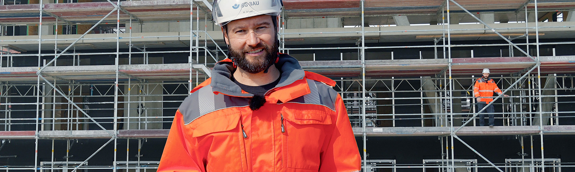 Eine Aufsichtsperson steht vor einem Gerüst. Im Hintergrund ist eine weitere Person auf einem Gerüst zu erkennen.