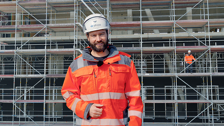 Eine Aufsichtsperson steht vor einem Gerüst. Im Hintergrund ist eine weitere Person auf einem Gerüst zu erkennen.