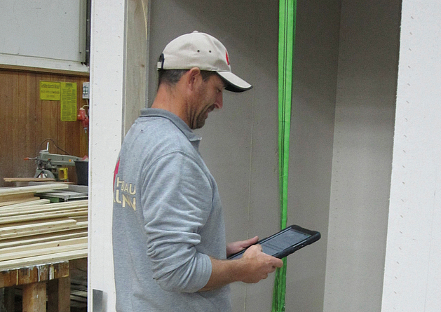 Mann in Werkhalle bedient Tablet, im Hintergrund liegen Holzbretter auf einem Werktisch.