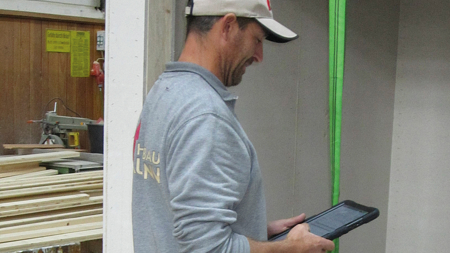 Dokumentation des betrieblichen AMS per Tablet Mann in Werkhalle bedient Tablet, im Hintergrund liegen Holzbretter auf einem Werktisch.