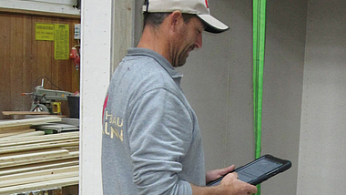 Mann in Werkhalle bedient Tablet, im Hintergrund liegen Holzbretter auf einem Werktisch.