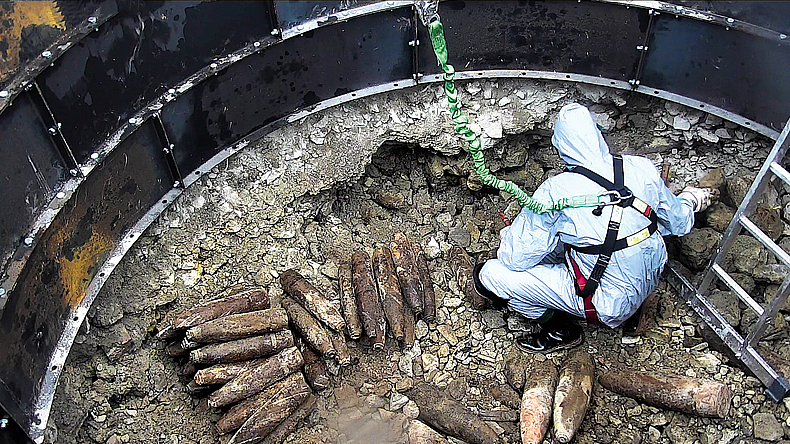 Ein Mann in Ganzkörperschutzanzug steht in einer Grube, in der Kampfmittel aneinandergereiht liegen.