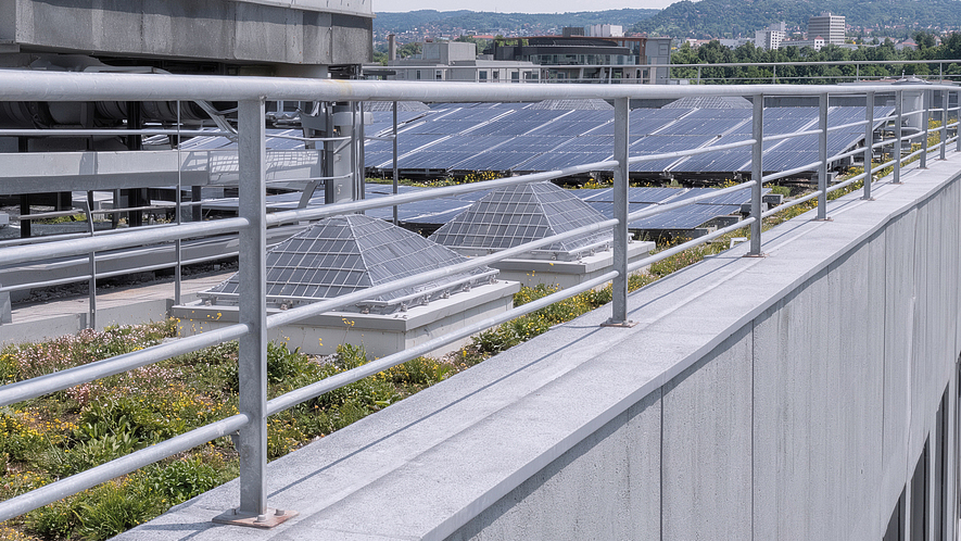 Flachdach mit mehreren Reihen von Solarmodulen, technischen Dachaufbauten und einem Metallgeländer im Vordergrund; im Hintergrund sind Gebäude und bewaldete Hügel zu sehen.