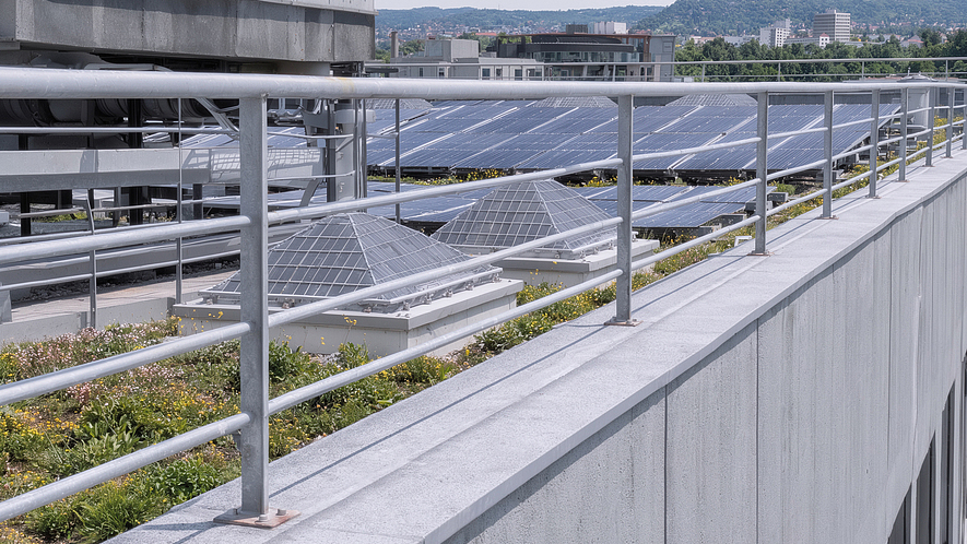 Flachdach mit mehreren Reihen von Solarmodulen, technischen Dachaufbauten und einem Metallgeländer im Vordergrund; im Hintergrund sind Gebäude und bewaldete Hügel zu sehen.