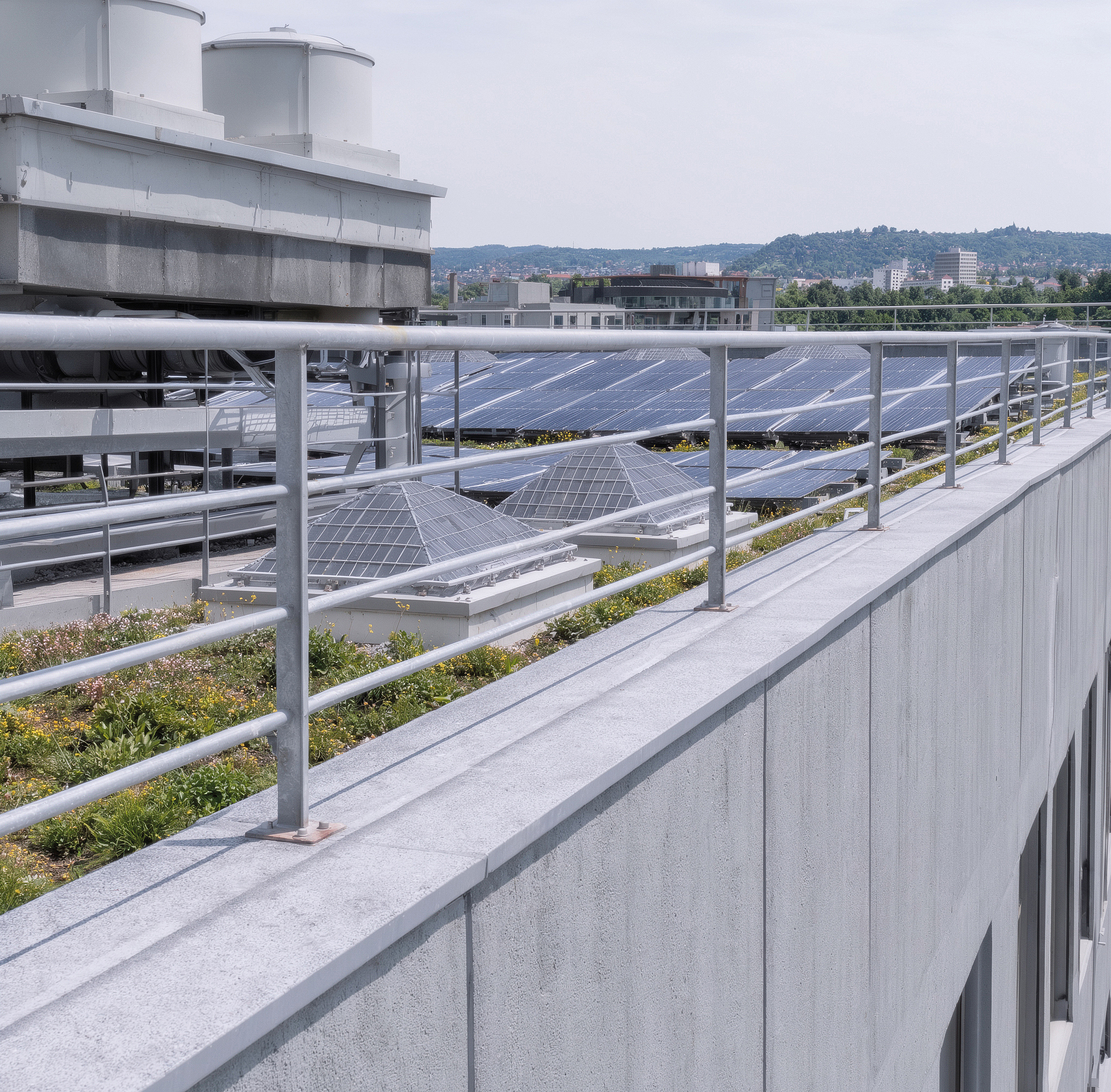 Flachdach mit mehreren Reihen von Solarmodulen, technischen Dachaufbauten und einem Metallgeländer im Vordergrund; im Hintergrund sind Gebäude und bewaldete Hügel zu sehen.