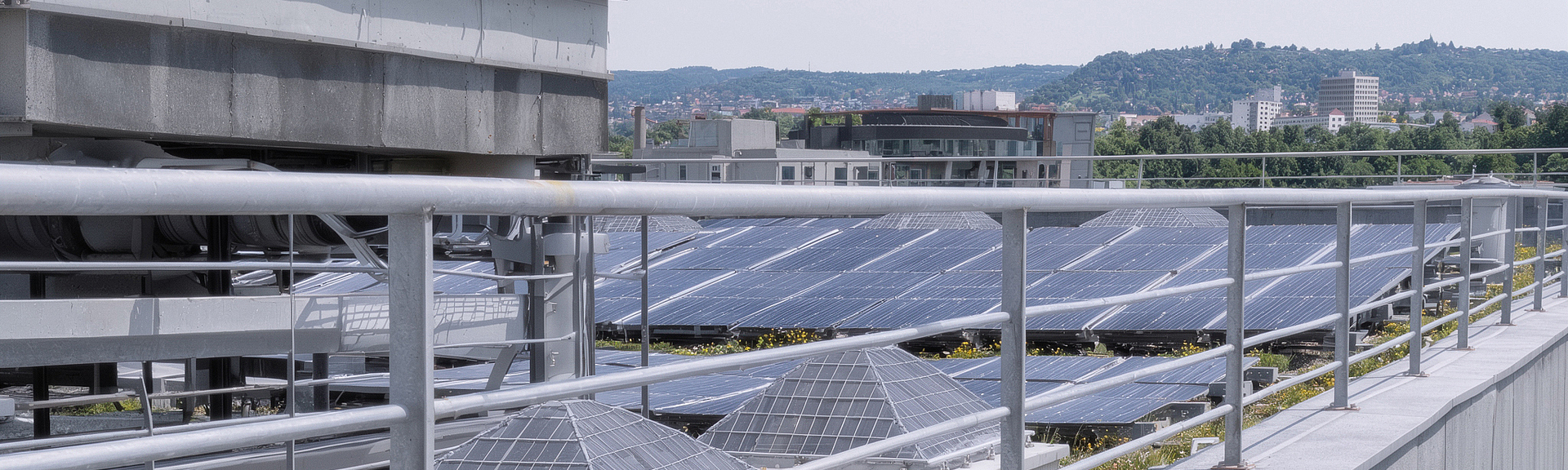 Flachdach mit mehreren Reihen von Solarmodulen, technischen Dachaufbauten und einem Metallgeländer im Vordergrund; im Hintergrund sind Gebäude und bewaldete Hügel zu sehen.