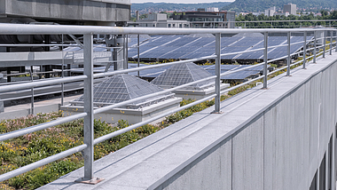 Flachdach mit mehreren Reihen von Solarmodulen, technischen Dachaufbauten und einem Metallgeländer im Vordergrund; im Hintergrund sind Gebäude und bewaldete Hügel zu sehen.