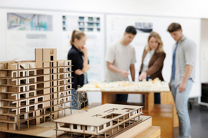Architekturmodelle aus Karton stehen im Vordergrund auf einem Tisch. Im unscharfen Hintergrund betrachten vier Personen gemeinsam ein weiteres Modell in einem Raum mit Plänen an den Wänden.