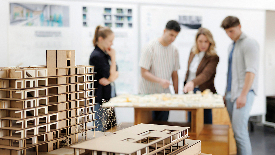 Architekturmodelle aus Karton stehen im Vordergrund auf einem Tisch. Im unscharfen Hintergrund betrachten vier Personen gemeinsam ein weiteres Modell in einem Raum mit Plänen an den Wänden.