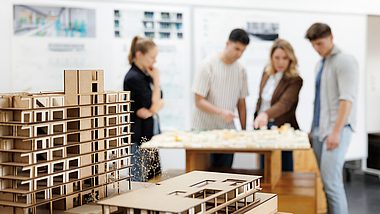 Zwei Architekturmodelle aus Pappkarton stehen im Vordergrund auf einem Tisch. Im unscharfen Hintergrund betrachten vier Personen gemeinsam ein weiteres Modell.