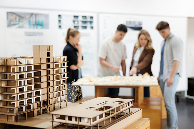 Zwei Architekturmodelle aus Pappkarton stehen im Vordergrund auf einem Tisch. Im unscharfen Hintergrund betrachten vier Personen gemeinsam ein weiteres Modell.