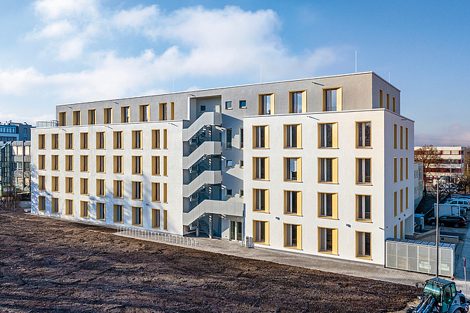 Außenansicht des kompakten Gebäudes: fünf Geschosse, zum Teil mit Außentreppen, und eine Dachterrasse.