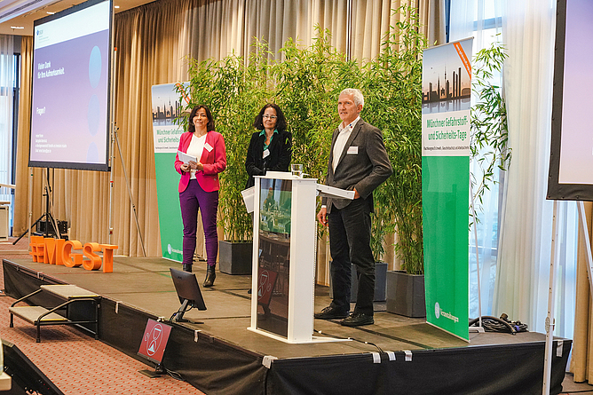 Zwei Frauen, eine links und eine in der Mitte stehend, rechts ein Mann am Stehpult auf einer Bühne.