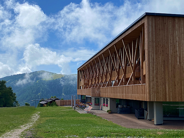 13 ineinandergreifende Holzstützen bilden die Kolonnade. Die gekreuzten Stämme sind aufgeschichtet und zu Verstrebungen verbunden. Sie tragen das zu Flügeln geformte neue Dach. Ein Gebäude mitten in den Alpen. Das Erdgeschoss ist aus Beton gebaut. Der obere Teil ist eine komplexe Holzkonstruktion.
