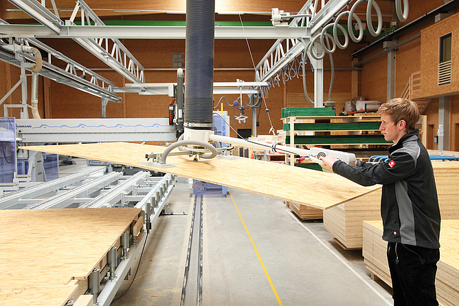 Mann bewegt in Werkhalle eine große Holzplatte mit einer Hebevorrichtung. Im Hintergrund sind Maschinen und gestapelte Holzplatten zu sehen.