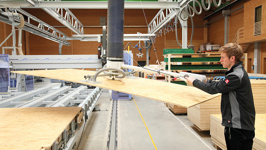 Mann bewegt in Werkhalle eine große Holzplatte mit einer Hebevorrichtung. Im Hintergrund sind Maschinen und gestapelte Holzplatten zu sehen.