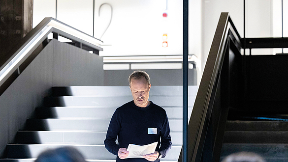 Mann steht in einem Treppenhaus und liest aus einem Blatt Papier vor.