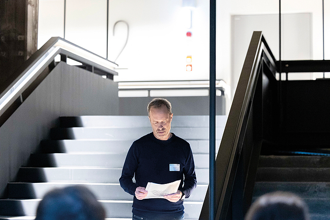Mann steht in einem Treppenhaus und liest aus einem Blatt Papier vor.