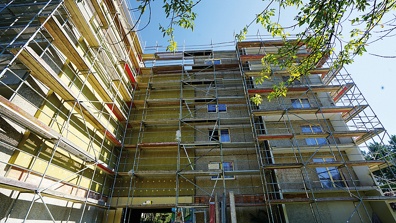 Eingerüsteter Plattenbau über Eck, ein Teil des Plattenbaus ist mit gelbgrünen Wärmedämmplatten versehen.