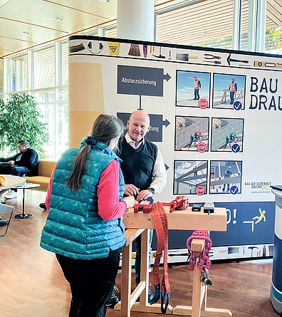 Ein Mann steht an einem Holzgestell vor einem großen BG-BAU-Poster und spricht mit einer Besucherin.