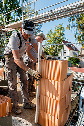 Zwei Männer in Arbeitskleidung stehen an einer etwa 80 cm hohen Wand aus großen Ziegelblöcken. Der linke Mann hat am Ende der Mauer eine Wasserwaage angelegt.