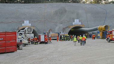 Baustelle mit zwei Tunnelportalen in einer Betonwand; davor stehen Einsatzkräfte und Bauarbeiter in Schutzkleidung neben Fahrzeugen und Geräten.