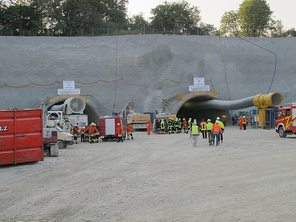 Baustelle mit zwei Tunnelportalen in einer Betonwand; davor stehen Einsatzkräfte und Bauarbeiter in Schutzkleidung neben Fahrzeugen und Geräten.