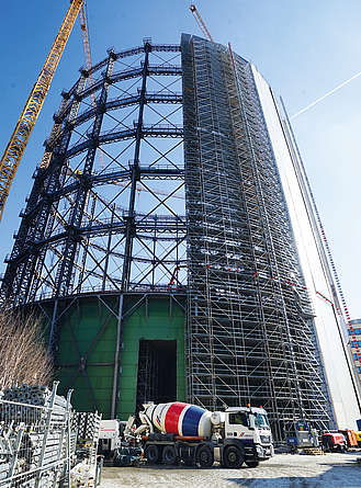 Blick auf das eingerüstete Gasometer, neben dem Bauwerk befindet sich ein Kran, davor ein Betonmischer.