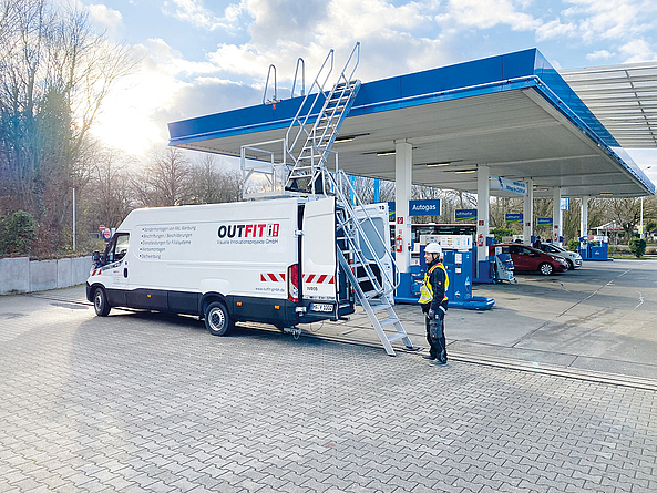 Zu sehen ist ein Transporter mit einer geöffneten Hintertür und zwei Leitern. An der geöffneten Tür steht eine Leiter, die vom Boden bis zum Dach des Transporters reicht. Auf dem Dach befindet sich eine weitere Leiter, die bis zum Tankstellendach reicht. Davor steht ein Mann in Schutzkleidung und Helm.