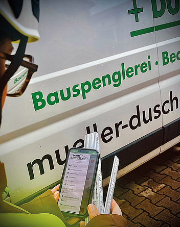 Person mit Helm hält ein Smartphone mit der geöffneten MÜLLER + DUSCHER App zur Gefährdungsbeurteilung vor einem Firmenfahrzeug, 