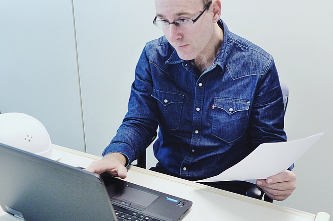 Ein Mann sitzt am Laptop mit Unterlagen in der Hand.