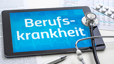 Auf einem Tisch liegt ein Tablet mit der Aufschrift Berufskrankheit. Darüber liegt ein Stethoskop.