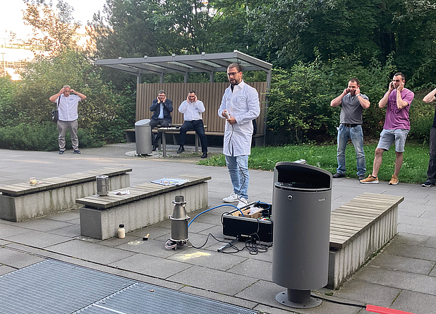 Ein Mann in weißem Kittel führt auf einem gepflasterten Platz ein Experiment durch. Mehrere Personen beobachten ihn aus sicherer Entfernung und halten sich die Ohren zu. Auf dem Boden stehen Laborgeräte und Behälter.