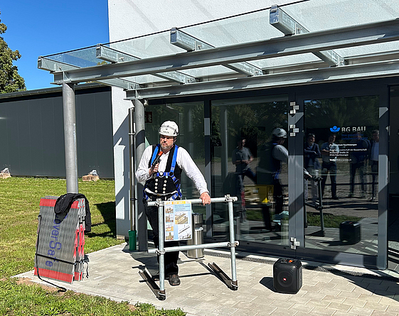 Ein Mann mit Schutzhelm und Auffanggurt demonstriert vor einem Gebäude der BG BAU eine Absturzsicherung an einem Gerüstmodell.
