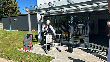 Ein Mann mit Schutzhelm und Auffanggurt demonstriert vor einem Gebäude der BG BAU eine Absturzsicherung an einem Gerüstmodell.