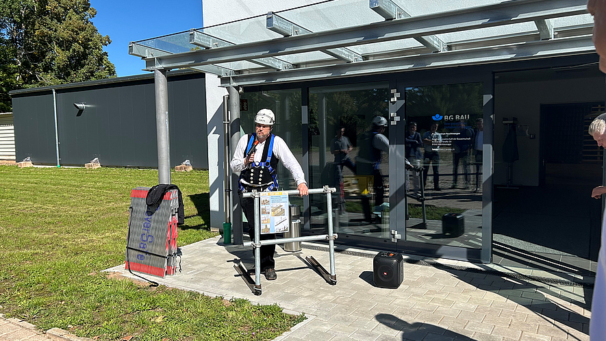 Elmar Mette mit Prototyp für den Seitenschutz Ein Mann mit Schutzhelm und Auffanggurt demonstriert vor einem Gebäude der BG BAU eine Absturzsicherung an einem Gerüstmodell.